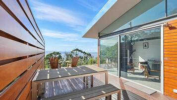 A modern outdoor deck with wooden furniture, a glass door leading to a balcony, and a view of the ocean.
