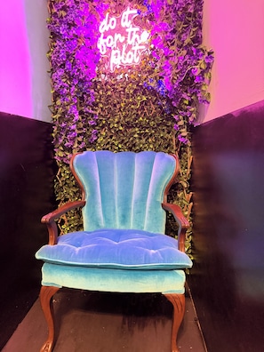 Un sillón tapizado en azul terciopelo con patas de madera, ubicado frente a una pared con plantas trepadoras y un letrero de neón que dice 'do it for the plot'.
