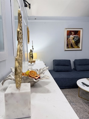 Un salon moderne avec un comptoir en marbre blanc, un bol décoratif en argent contenant des fruits secs, un canapé bleu foncé et un tableau encadré accroché au mur.