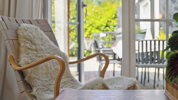 A wicker chair with a fluffy white cushion in a sunlit room with a view of outdoor furniture and greenery. - Cozy holiday home in scenic Rågeleje (Vejby)