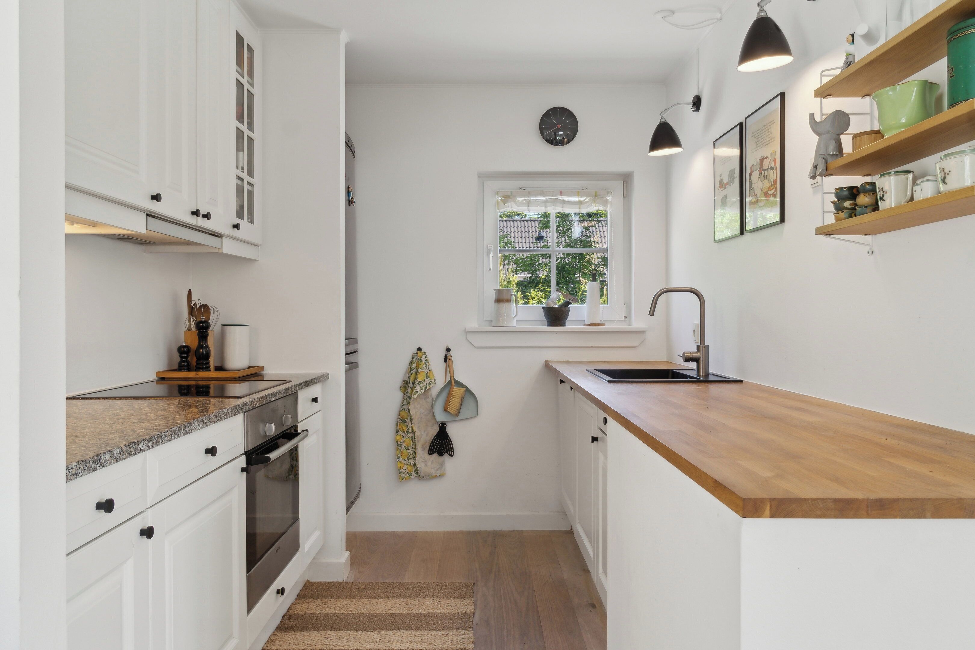Cocina compacta con gabinetes blancos, encimera de madera, horno empotrado, fregadero y ventana con vistas a zonas verdes.