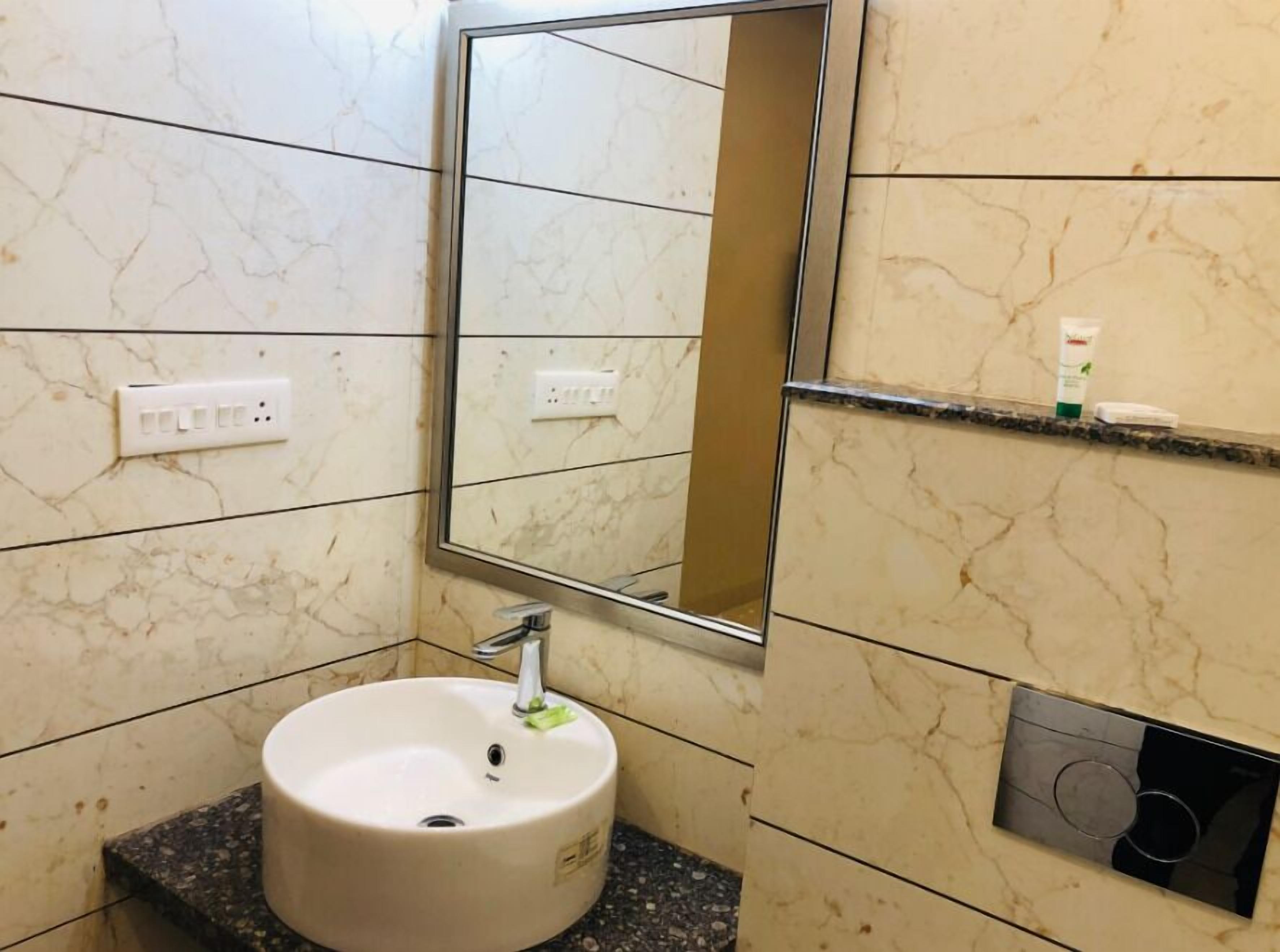 A bathroom with a marble wall, a sink, a mirror, a shelf with toiletries, and a flush toilet.
