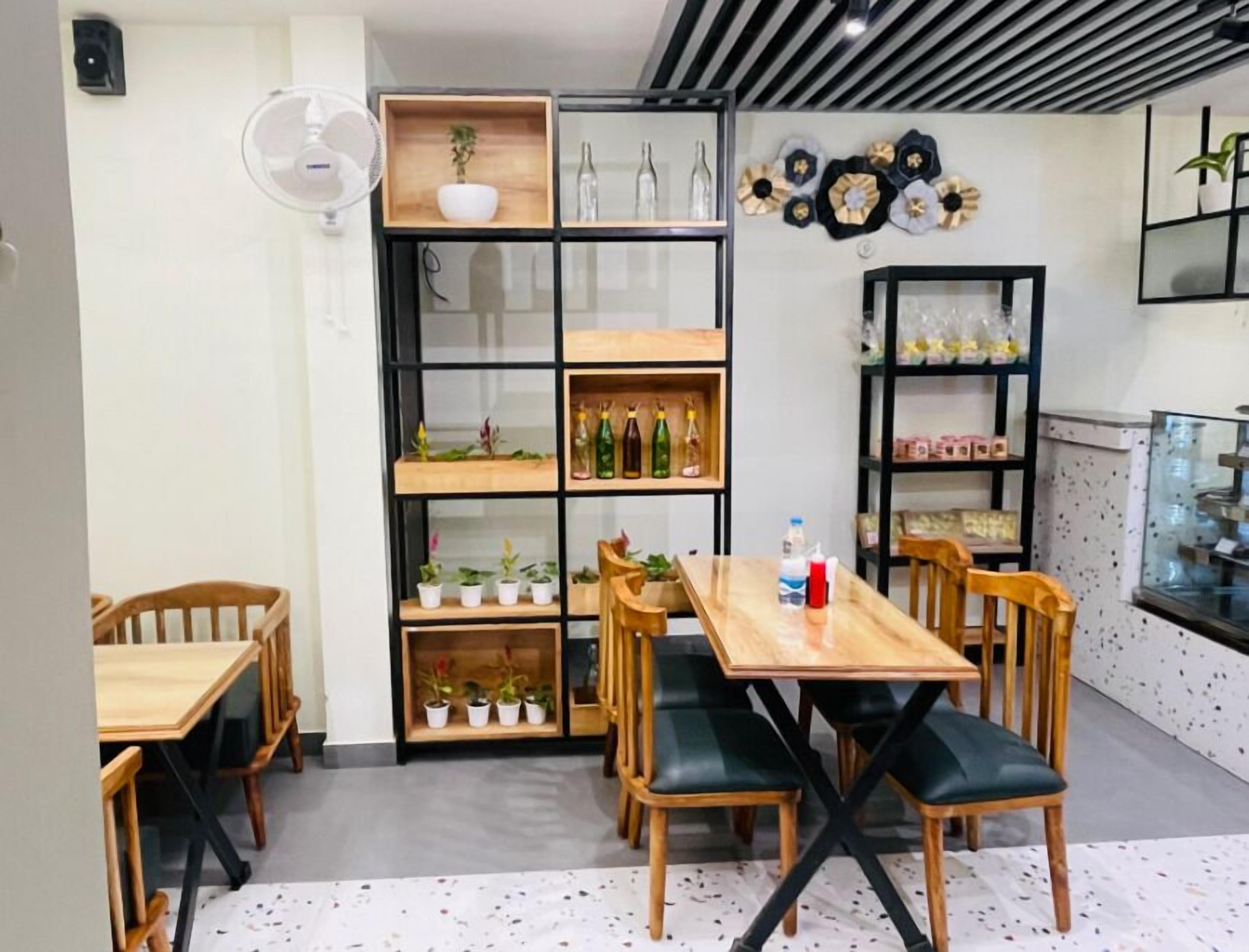 A dining area with wooden tables and chairs, a central table with a bottle, and shelves with plants and bottles.