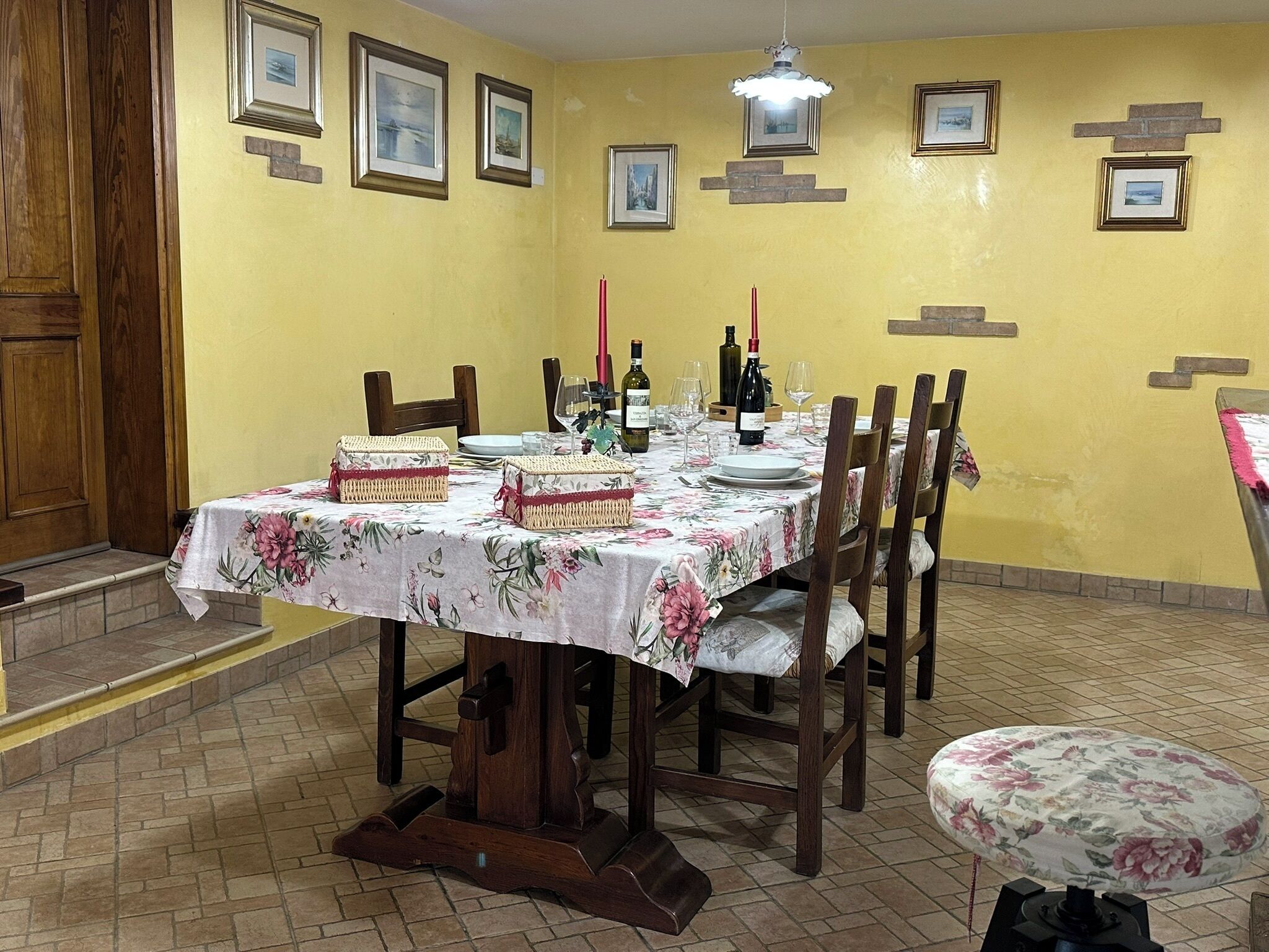 Un coin repas avec une table dressée pour quatre personnes, des chaises en bois, une nappe à motifs floraux et des tableaux encadrés accrochés au mur.
