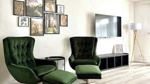 A modern living room with two green velvet armchairs, a flat-screen TV, and a wall decorated with framed pictures.