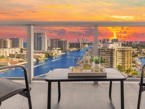A balcony with a table set for two, overlooking a cityscape and a river at sunset.