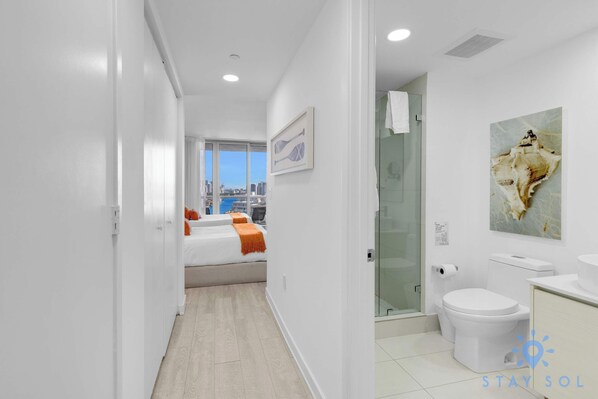 A modern hotel room with a city view, a bed with orange bedding, a glass shower, and a white toilet.