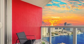 A balcony with a red wall, a black chair, and a view of a cityscape and water at sunset.
