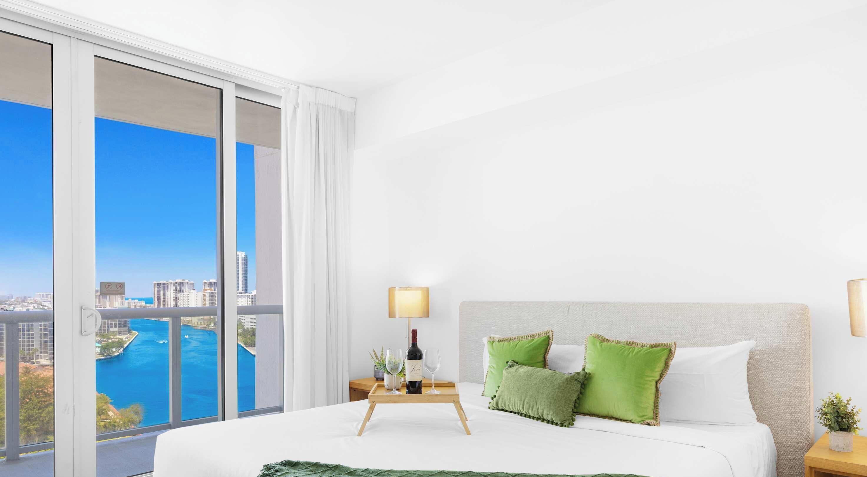 A hotel room with a bed, a nightstand, a lamp, and a view of a cityscape and water.
