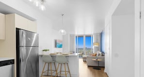 Cozinha moderna com geladeira de aço inoxidável, dois bancos de bar, área de jantar com mesa e cadeiras, sofá e vista da cidade por grandes janelas.