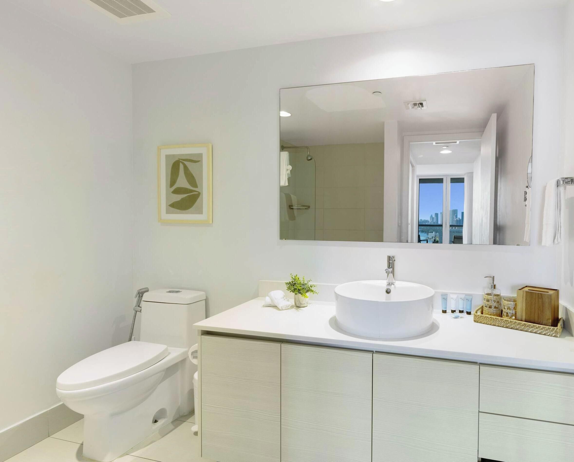 A modern bathroom with a large mirror, a round sink, a toilet, and a framed artwork on the wall.