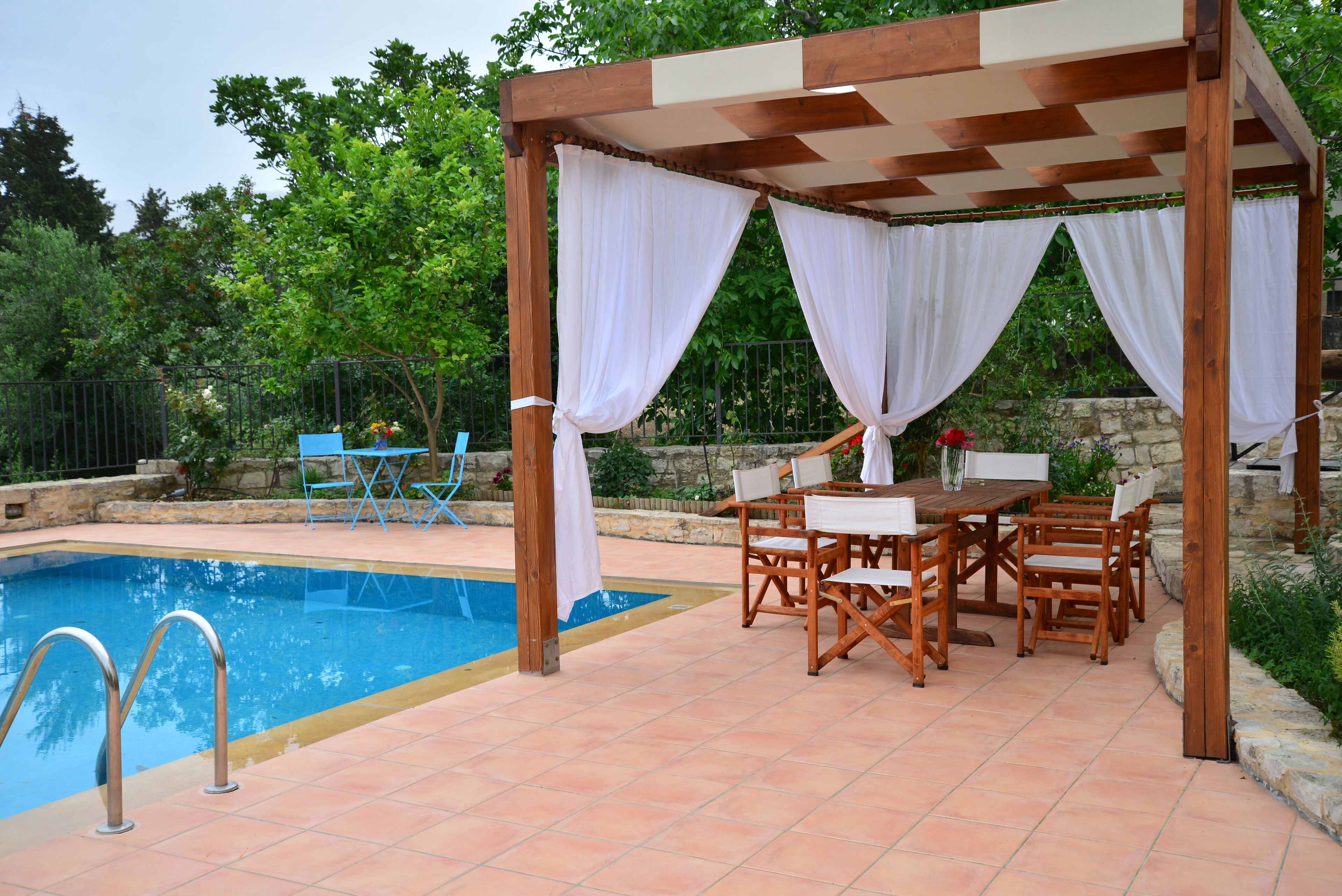 Zona junto a la piscina con un porche de madera, cortinas blancas y una mesa con sillas.