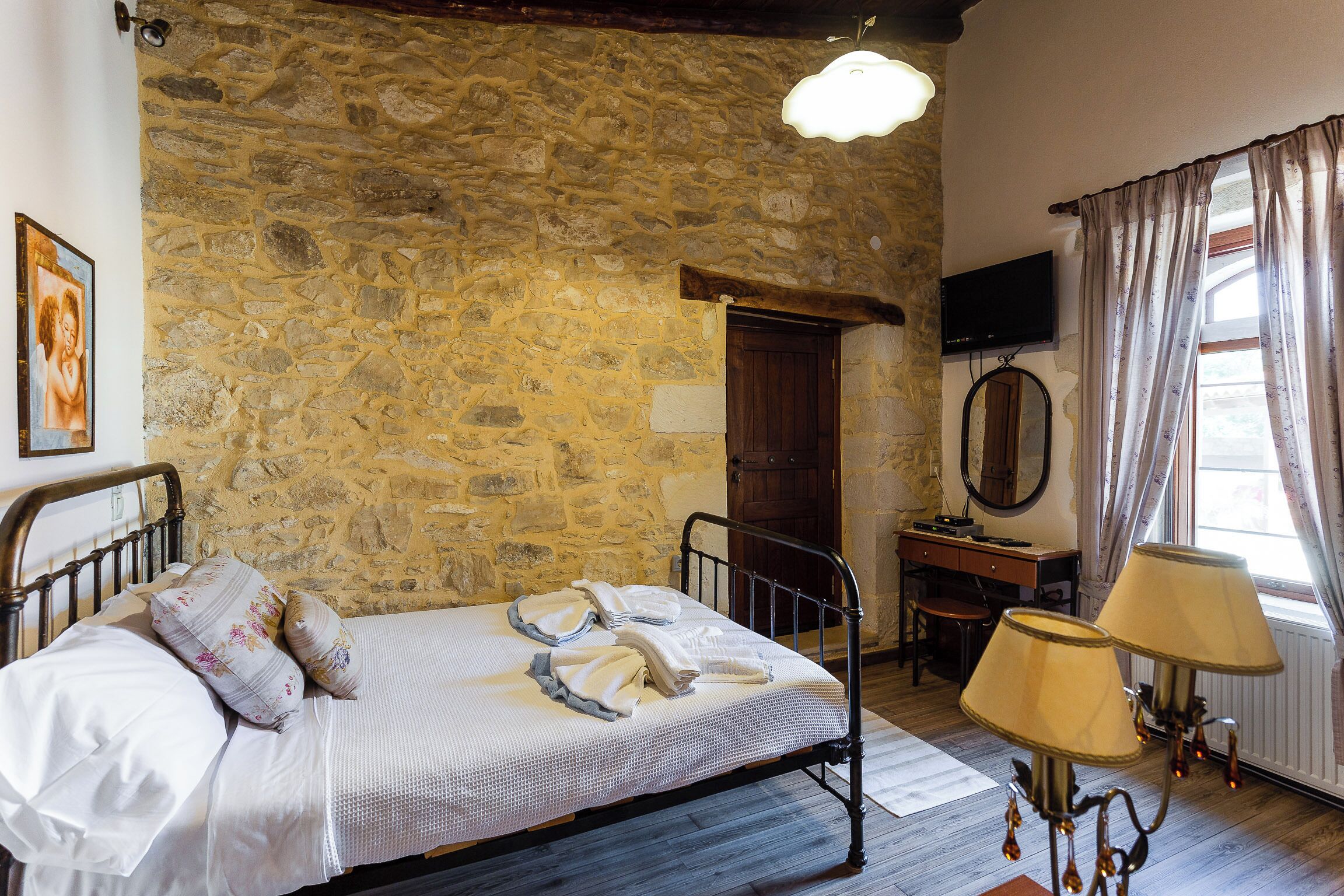 A bedroom with a stone wall, a bed with white linens, a wooden desk, a television, and a window with curtains.