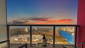 A view from a high-rise window overlooking a cityscape with a river, a sunset, and a bottle of wine on a table. - Amazing 2BR w/ Private Balcony + Views + Pool (Hallandale Beach)
