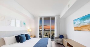 A hotel room with a bed, a desk, a chair, a painting, and a view of the sunset through large windows. - Amazing 2BR w/ Private Balcony + Views + Pool (Hallandale Beach)