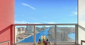 A balcony with a view of a river and cityscape, a table with bottles and grapes, and a red wall.