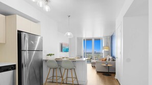 A modern kitchen with a stainless steel refrigerator, two bar stools, a dining area with a table and chairs, a sofa, and a view of the city through large windows. - Amazing Apt Private Balcony + Pool + Amenities (Hallandale Beach)