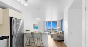 Cozinha moderna com geladeira de aço inoxidável, dois bancos de bar, área de jantar com mesa e cadeiras, sofá e vista da cidade por grandes janelas.