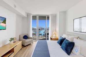 A hotel room with a bed, a desk, a chair, a view of the city, and a balcony with a table and chairs.