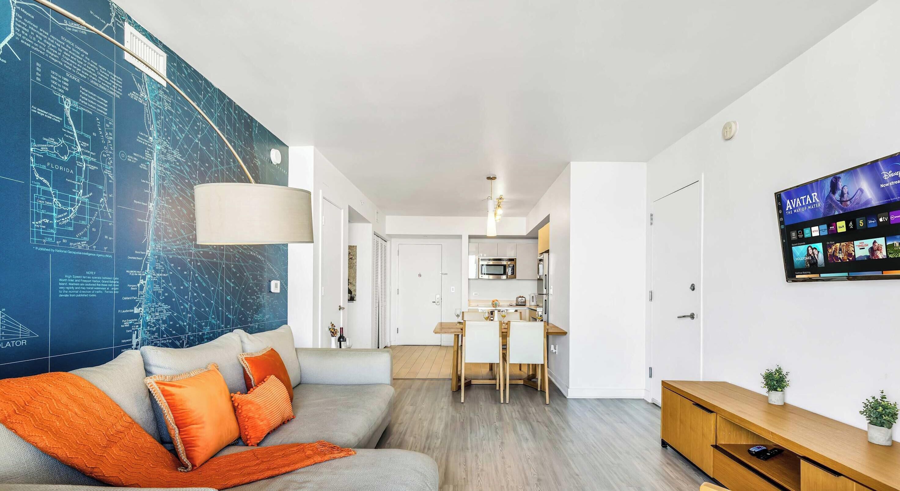 Ein modernes Wohnzimmer mit einer großen Karte an der Wand, einem Sofa mit orangen Kissen, einem Essbereich und einem Fernseher, der einen Film zeigt.