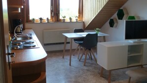 Eine Küche mit Herd, Spüle und Fenster mit Pflanzen. Es gibt einen Esstisch mit Stühlen und einen Fernseher.