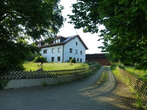 A two-story house with a sloped roof, surrounded by greenery and a paved driveway.