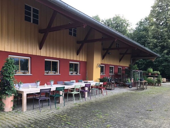 A building with a red and yellow exterior, a covered outdoor seating area with tables and chairs, and a garden with potted plants.