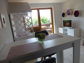 Quarto com mesa de jantar, televisão em móvel, cadeira, uma pequena planta e varanda com guarda-corpo de madeira.