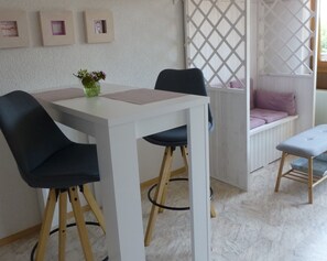 Une pièce avec une table blanche, deux chaises noires, un petit vase vert, un banc blanc avec un coussin violet et un tabouret en bois.