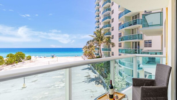 A balcony with a clear view of the beach and ocean, a dining table with a bottle and glasses, and a wicker chair. - Beautiful Apartment with Pool & Amenities (Hollywood)