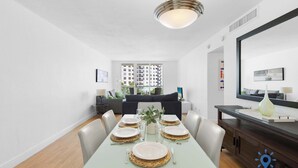 A dining area with a glass table set for four, a dark wooden sideboard, and a large mirror above the table. - Beautiful Apartment with Pool & Amenities (Hollywood)