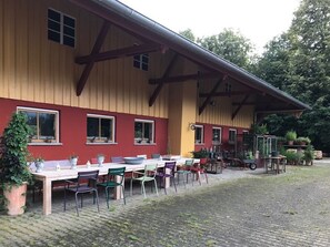 Een gebouw met een rode en gele gevel, een overdekt terras met tafels en stoelen, en een tuin met planten in potten.