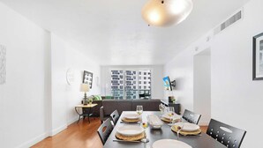 Un área de comedor moderna con una mesa de madera dispuesta para una comida, un sofá y vistas a la ciudad a través de amplias ventanas.