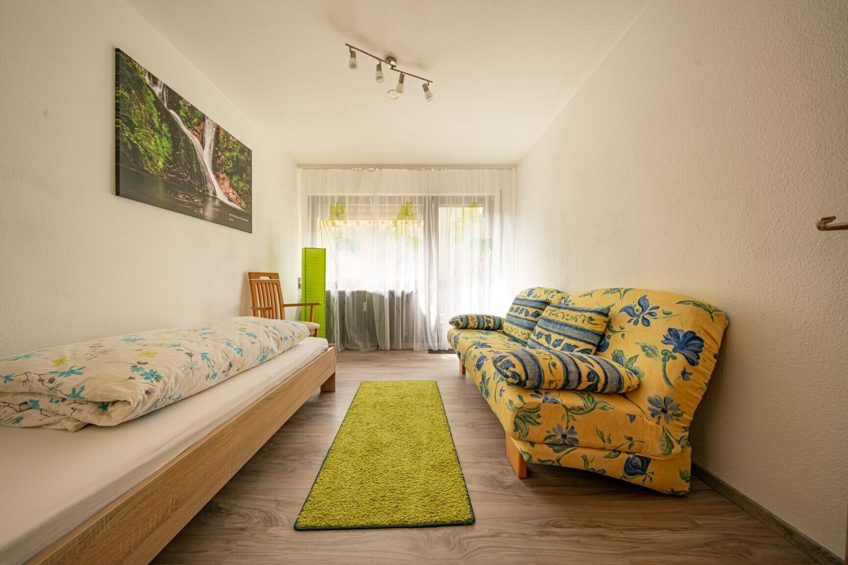 Un dormitorio con cama, sofá, alfombra verde, ventana con cortinas e imagen colgada en la pared.