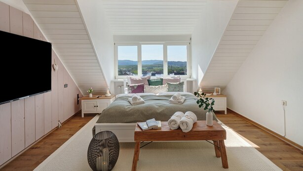 A bedroom with a bed, a bedside table, a TV, a window with a view, and a wooden ceiling.