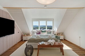 A bedroom with a bed, a bedside table, a TV, a window with a view, and a wooden ceiling.