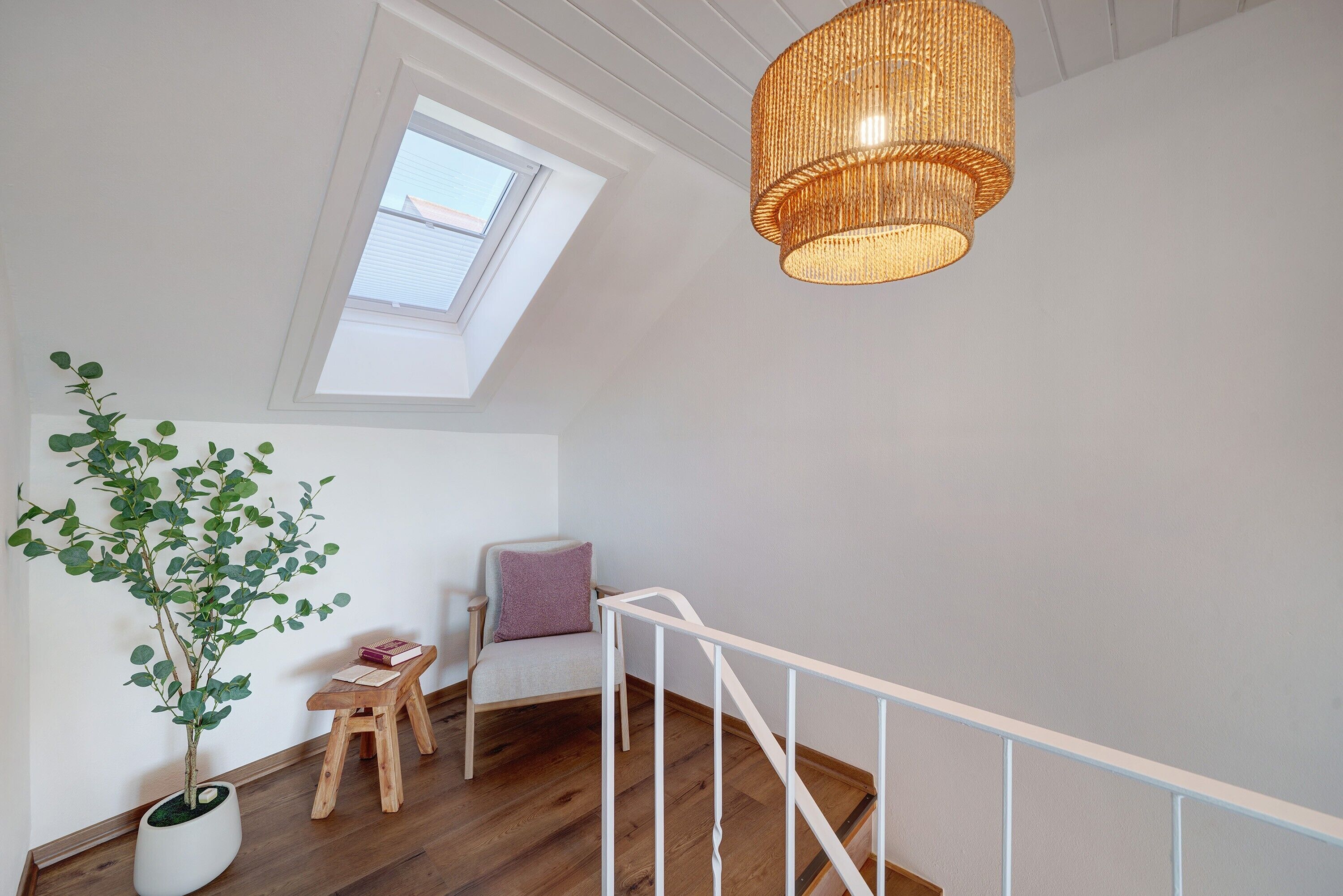 Ett rum med trägolv, ett vitt räcke, en krukväxt, en stol med en kudde och en hängande lampa.