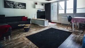 A modern living room with a black sofa, a flat-screen TV, a dining table with a checkered tablecloth, and a large window with blinds. - Apartment 'Natur Tanken' with Mountain View, Shared Garden and Wi-Fi (Sasbachwalden)