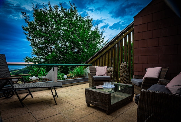 An outdoor patio with wicker furniture, a glass table, and a view of the sky and greenery.
