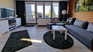 A modern living room with a flat-screen TV, a sofa, a coffee table, and a large window with a view of greenery.
