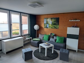 Un salón moderno con un sofá gris, una mesa de centro blanca, una alfombra redonda y un cuadro de un bosque en la pared.