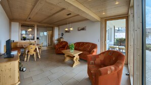 A spacious living area with a wooden ceiling, a dining table with chairs, a sofa, and a large window offering a view.