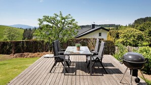 A wooden deck with a table and chairs, a barbecue grill, and a house in the background.