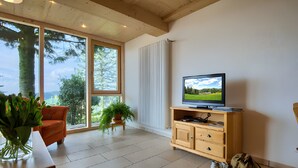 A room with a television, wooden furniture, a potted plant, and a view of trees through large windows.
