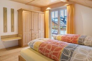 A bedroom with two beds, a wooden wardrobe, a small shelf, and a large window with curtains.