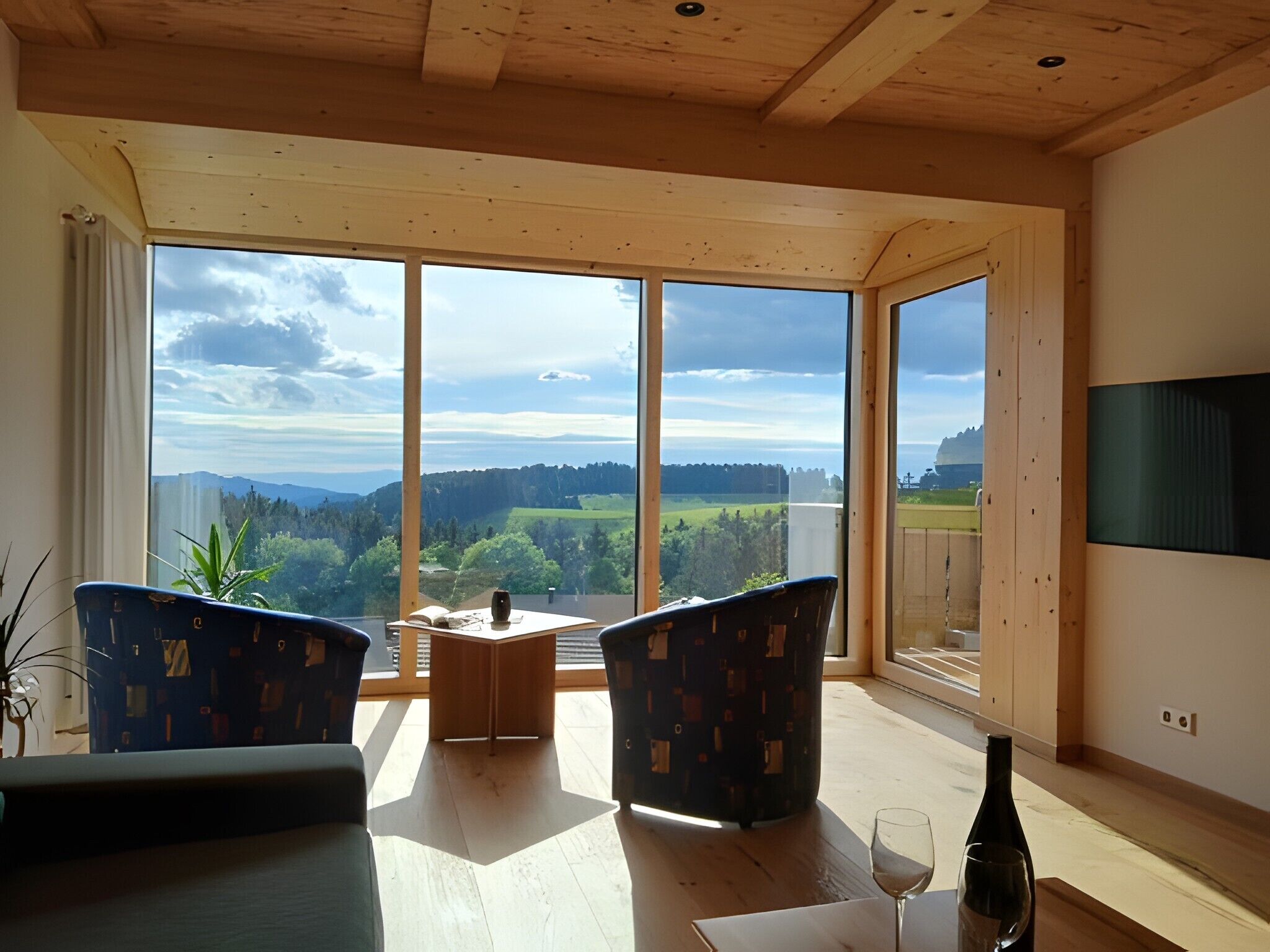 A room with a large window offering a view of a landscape, two chairs, a small table, and a bottle of wine.