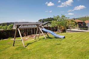 Une aire de jeux avec un toboggan et une balançoire, entourée d’une cour verdoyante et d’une maison avec un toit rouge.