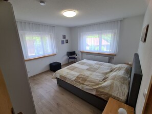 Une chambre à coucher avec un lit, deux fenêtres avec des rideaux, un radiateur et un petit tabouret.