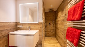 Un baño moderno con lavamanos blanco, paredes de madera, espejo con iluminación integrada, zona de ducha y toallas rojas.