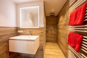 A modern bathroom with a white sink, wooden walls, a mirror with integrated lighting, a shower area, and red towels.
